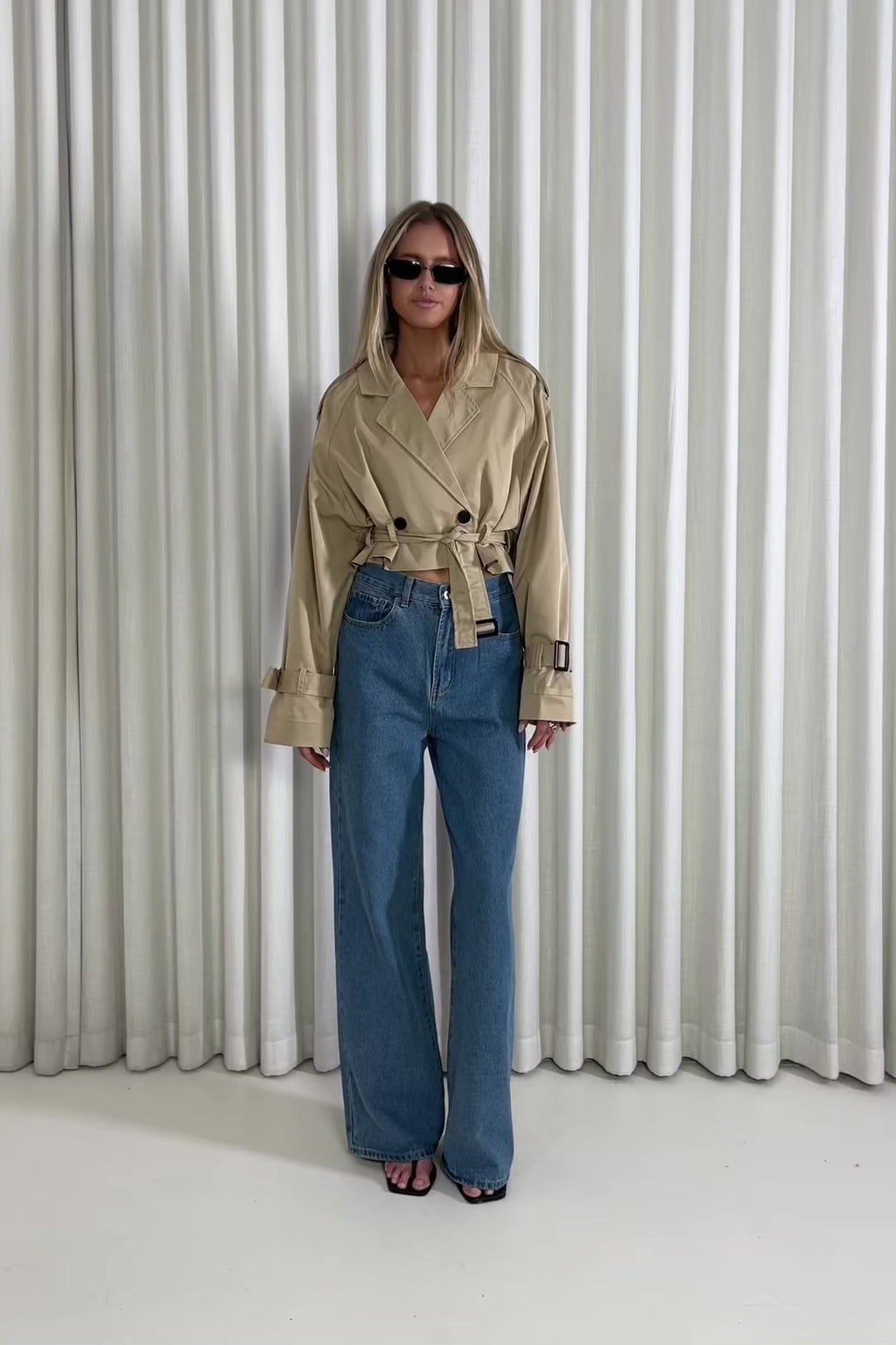 Woman wearing a cropped beige trench and denim jeans against a white curtain backgrounds