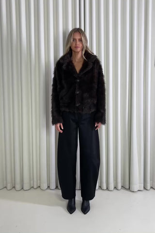 Woman wearing a brown jacket and black pants standing against a white curtain, showing of the outfit.