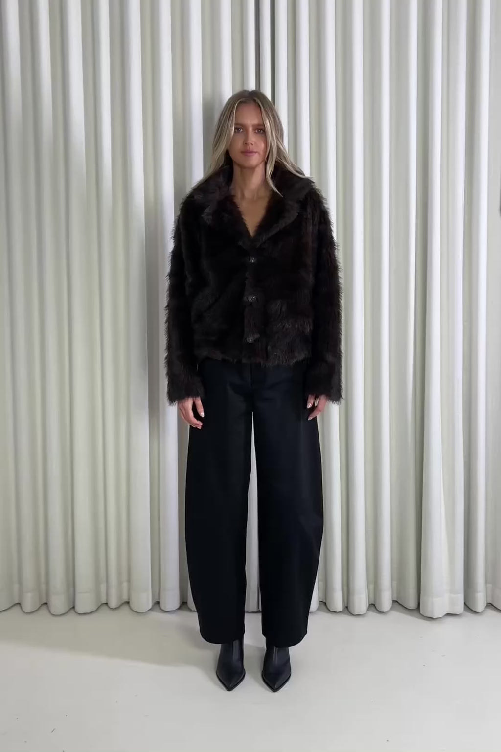 Woman wearing a brown jacket and black pants standing against a white curtain, showing of the outfit.