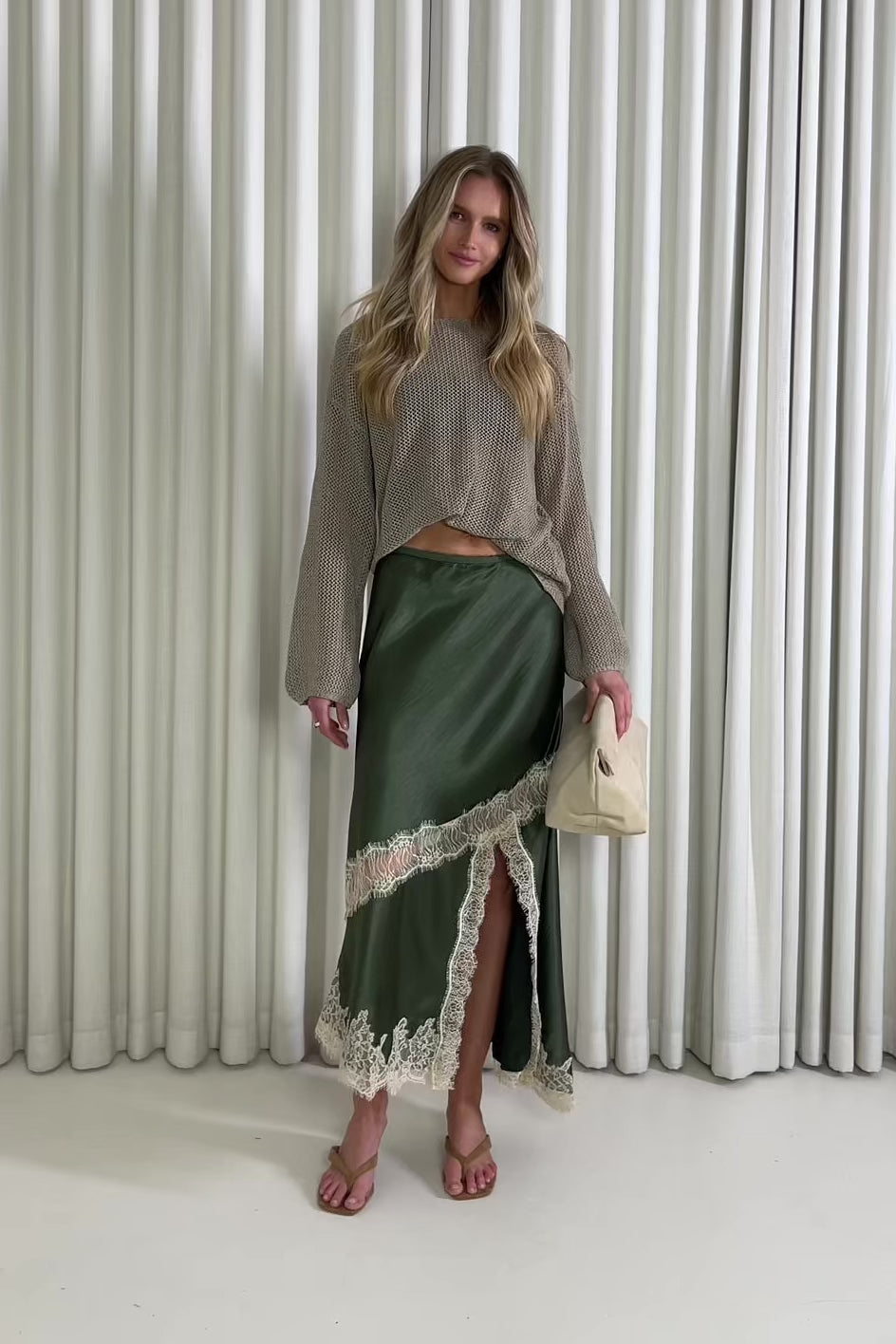 Woman wearing a green satin skirt with lace trim and a beige sweater against a white curtain background.