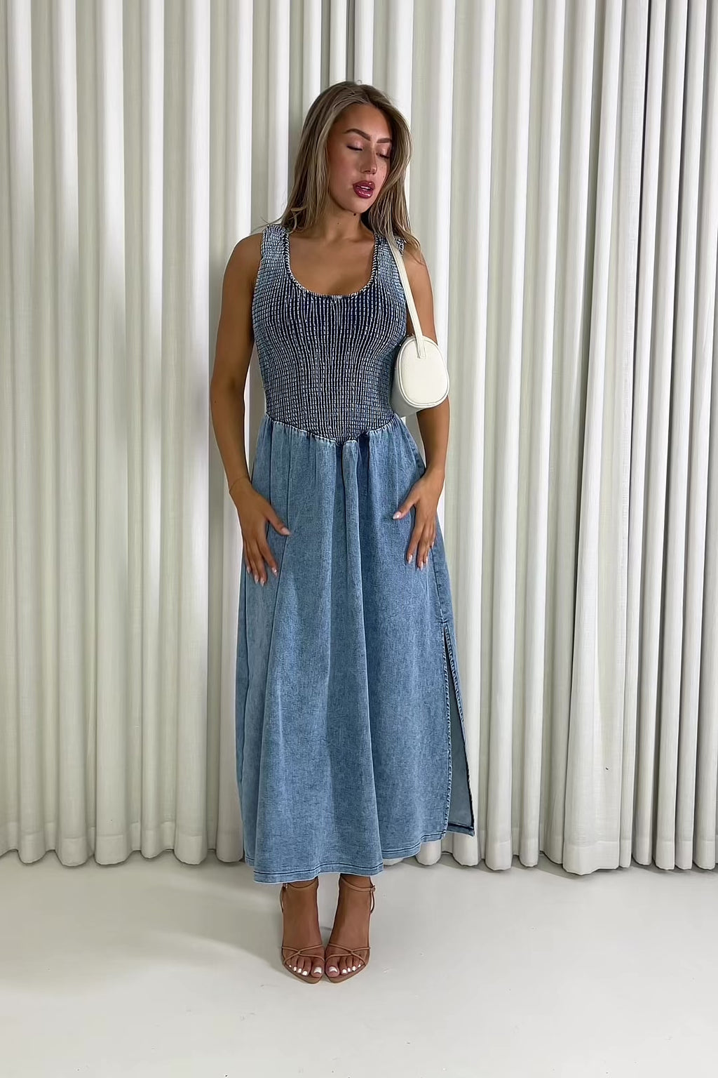 Woman wearing a blue dress holding a white clutch against a white curtain background Eddie denim midi dress with shirred bust, racerback
