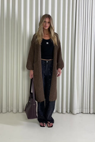 Person wearing a long brown cardigan over a black top and jeans, standing against a white curtain background.