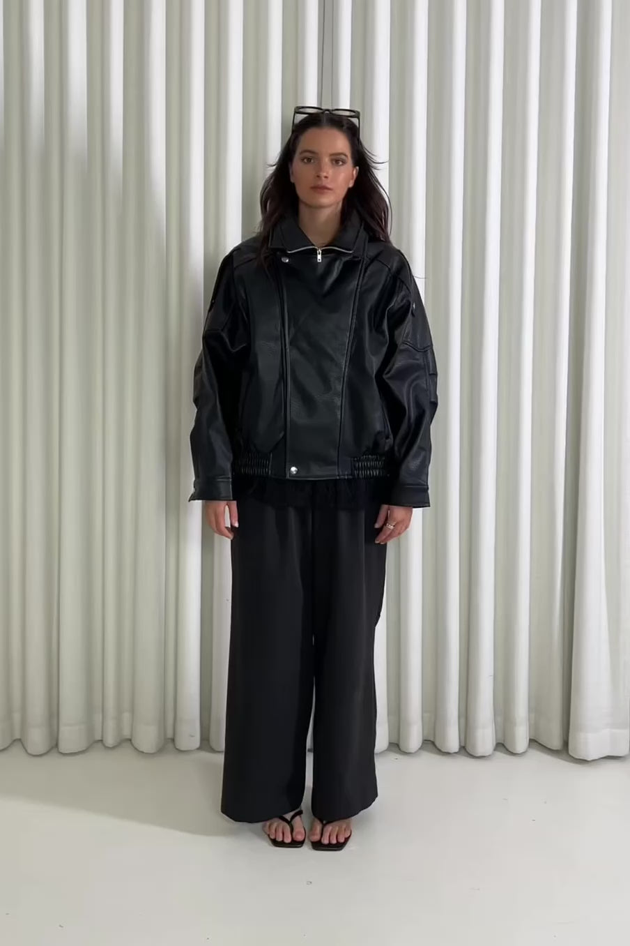Person wearing a black oversized faux leather bomber and pants standing against a white curtain.