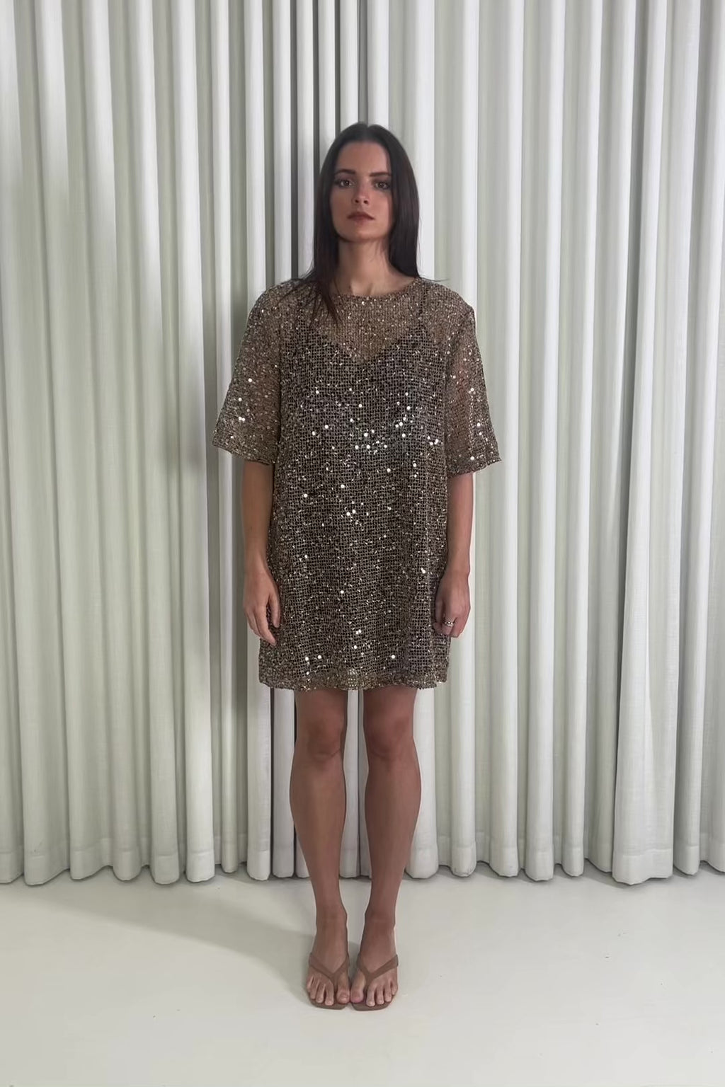 Woman wearing a sequin sheer dress standing against a white curtain.