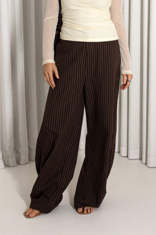A person wearing loose, wide-leg brown pants with thin vertical white pinstripes, paired with a cream long-sleeve top, standing barefoot in front of light gray curtains.