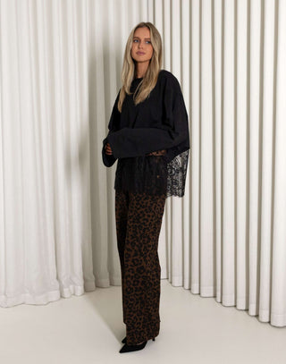 Woman wearing a black lace top and leopard print pants standing against a white curtain.