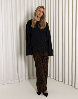 Woman wearing a black lace top and leopard print pants standing against a white curtain.