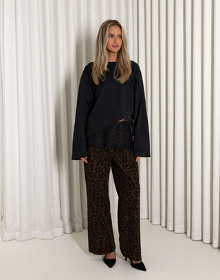 Woman wearing a black lace top and leopard print pants standing against a white curtain.