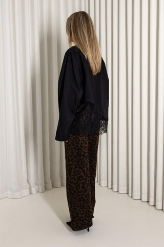 A woman stands with her back to the camera wearing a black long-sleeve top with lace trim and brown leopard-print pants, posed in front of white pleated curtains.