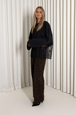 A woman modeling a black long-sleeve top with a sheer lace hem and wide-leg brown leopard-print pants, standing in front of a white pleated curtain backdrop.