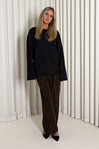 A woman modeling a black long-sleeve top with a sheer black lace overlay and brown leopard-print pants, standing in front of white pleated curtains.