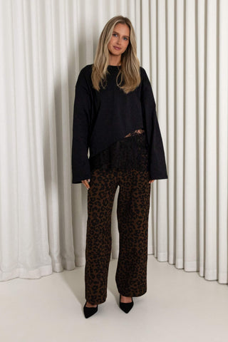 A woman stands in front of white pleated curtains wearing a black long-sleeve top with a sheer black lace overlay and wide-leg brown leopard-print pants, paired with black pointed-toe heels.