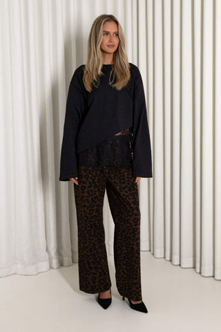 A woman stands against a white curtain backdrop wearing a black asymmetrical long-sleeve top with sheer lace detailing and wide-leg brown leopard-print pants.