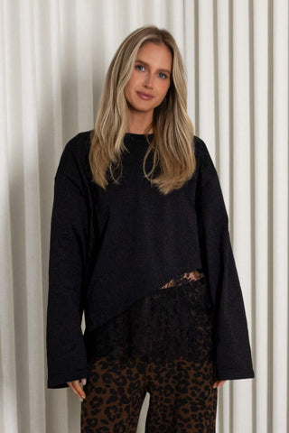 A woman with blonde hair wearing a black long-sleeve knit top with a lace hem detail and leopard print pants, standing in front of a white curtain background.