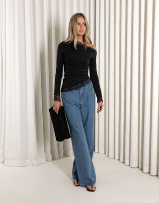 Woman wearing a black top and blue jeans standing against a white curtain.