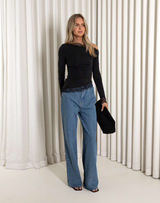 Woman wearing a black top and blue jeans holding a black clutch against a white curtain background