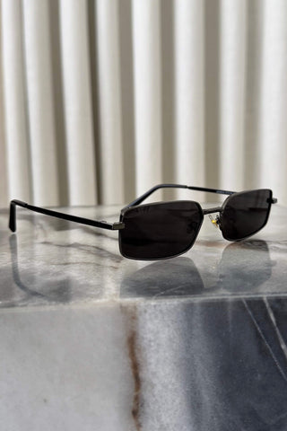 A pair of rimless square sunglasses with dark lenses and thin black metal frames, resting on a polished gray marble surface with a cream pleated curtain background.