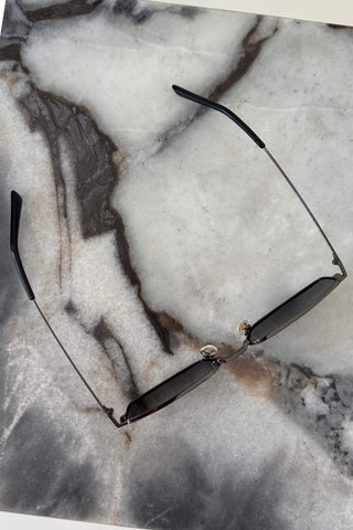 Thin metal-framed sunglasses with dark lenses and black temple tips, laid flat on a gray and white marble surface.