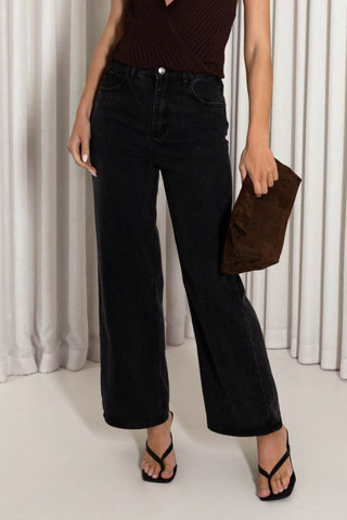 A model wearing black wide-leg cropped jeans, a dark brown ribbed knit top, and black thong sandals, holding a brown suede clutch against a background of white draped curtains.