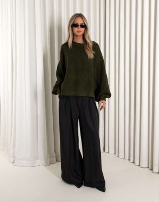 Person wearing a green sweater and black pants against a white curtain background