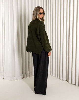 Person wearing a green jacket and black pants standing against a white curtain.