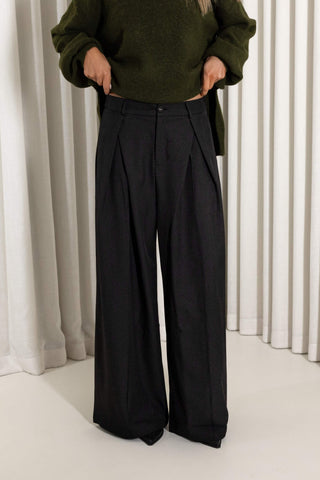 A person wearing black wide-leg pleated trousers and an olive green sweater, standing in front of white vertical drapes.