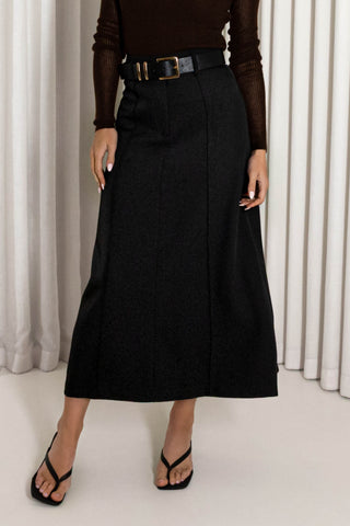 A model wearing a black wool-like A-line midi skirt with a black belt, paired with a dark brown sheer long-sleeve top and black thong sandals, set against a background of white vertical drapes.