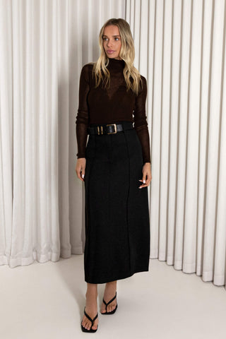 A woman modeling a dark brown sheer long-sleeve turtleneck top paired with a black midi skirt and black strappy sandals, standing in front of white pleated curtains.