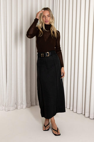 A woman stands in front of white draped curtains wearing a dark brown sheer long-sleeve top, a black high-waisted skirt with a black belt, and black sandals.