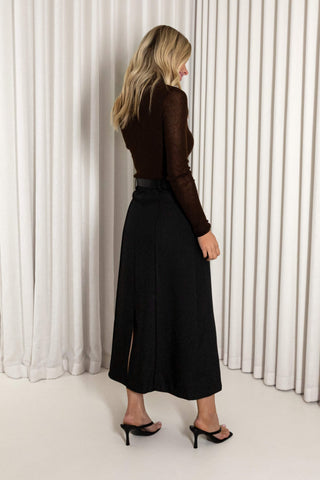A woman stands in profile wearing a dark brown long-sleeve knit top and a black midi skirt with a side slit, paired with black heeled sandals, set against a background of white vertical drapes.