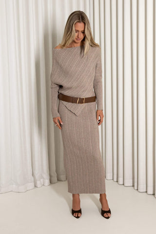 A woman models a ribbed knit off-the-shoulder sweater and matching midi skirt set in a taupe-beige color, styled with a brown belt and black heeled sandals against a white curtain backdrop.