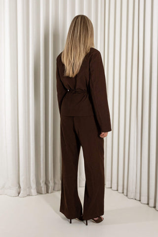 A woman modeling a dark brown belted wrap jacket and matching wide-leg trousers, standing against a white pleated curtain backdrop.