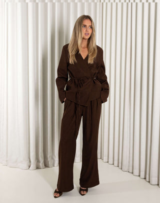 Woman wearing a brown blazer and brown matching pants standing against a white curtain background