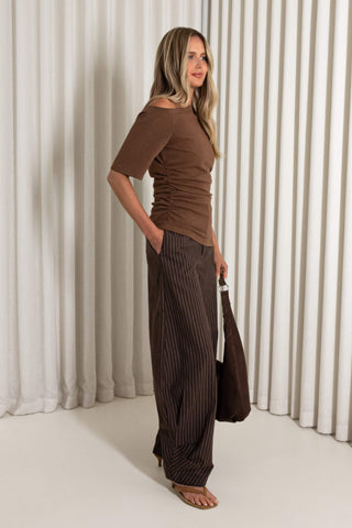 A woman modeling a brown off-the-shoulder short-sleeve top and matching brown pinstriped wide-leg pants, holding a dark brown tote bag against a background of vertical white drapes.