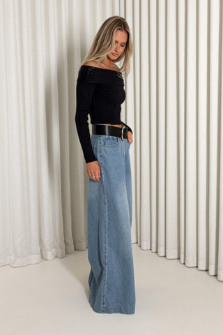 A woman stands in profile wearing a black off-the-shoulder long-sleeve top and wide-leg light blue denim jeans with a black belt, posed in front of a white pleated curtain backdrop.