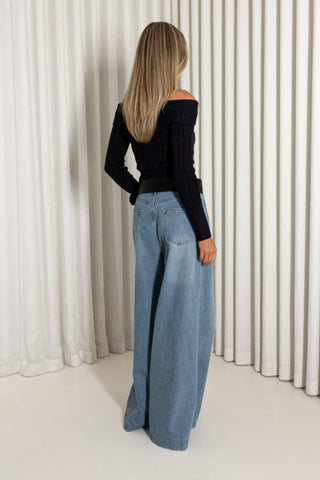 A woman with long blonde hair is shown from behind wearing a black off-the-shoulder ribbed long-sleeve top and wide-leg light blue denim jeans, standing in front of white vertical drapes.