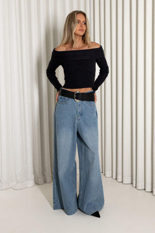 A woman modeling a black off-the-shoulder ribbed long-sleeve crop top and wide-leg light blue jeans with a black belt, standing in front of a white pleated curtain backdrop.