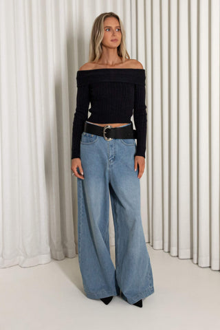 A woman modeling an off-the-shoulder black ribbed long-sleeve top paired with wide-leg light blue jeans and a black belt, standing against a white pleated curtain backdrop.
