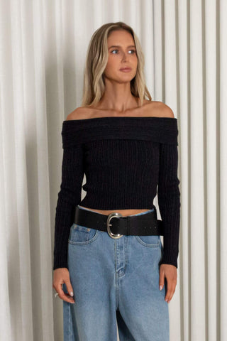 A woman modeling a black off-the-shoulder ribbed knit long-sleeve crop top paired with light blue jeans and a black belt, standing against a white curtain backdrop.