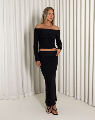Woman wearing a black off-shoulder top and black skirt against a white curtain background