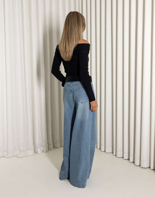 Woman wearing a black off-shoulder top and blue wide-leg jeans against a white curtain background