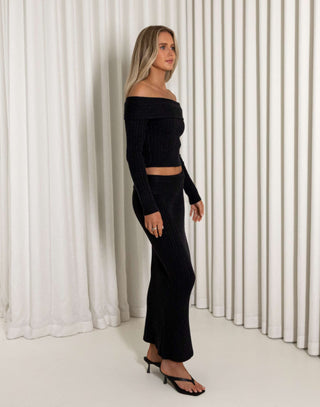 Woman wearing a black off-shoulder top and black skirt against a white curtain background