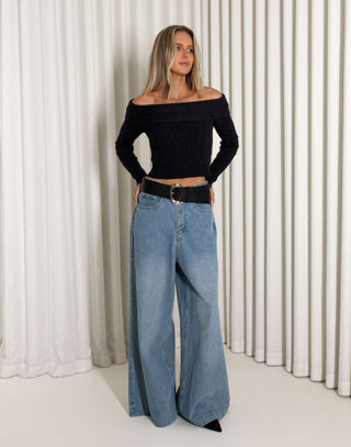 Woman wearing a black off-shoulder top and blue wide-leg jeans against a white curtain background