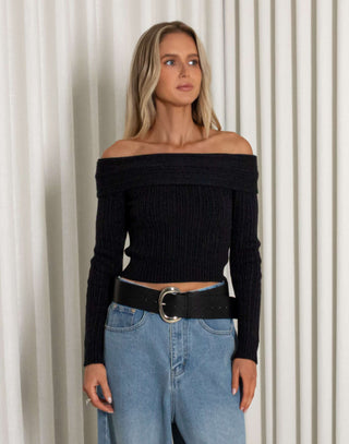 Woman wearing a black off-shoulder top and blue wide-leg jeans against a white curtain background