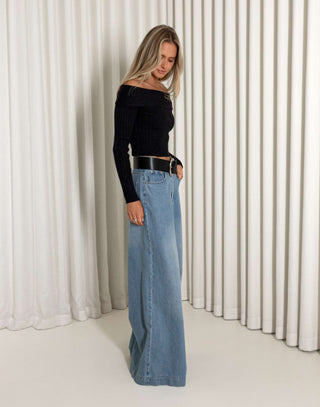Woman wearing a black off-shoulder top and blue wide-leg jeans against a white curtain background