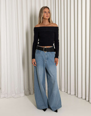 Woman wearing a black off-shoulder top and blue wide-leg jeans against a white curtain background