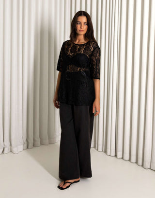 Woman wearing a black lace top and wide-leg pants against a white curtain background Black sheer lace longline t shirt style top