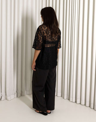 Woman wearing a black lace top and wide-leg pants against a white curtain background Black sheer lace longline t shirt style top