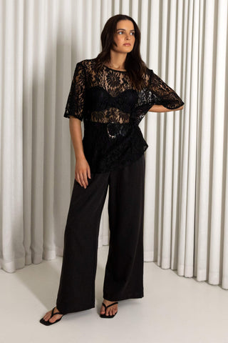 A woman modeling a black sheer lace short-sleeve top with a sweetheart neckline and black wide-leg pants, standing in front of white pleated curtains.