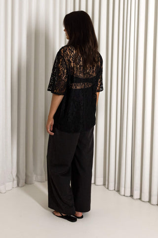 A woman viewed from behind wearing a black lace short-sleeve top and wide-leg black pants, standing in front of white pleated curtains.